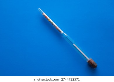 ​A glass hydrometer with a colored scale is positioned diagonally on a deep blue background. This laboratory device provides accurate measurement of liquid density and alcohol strength,