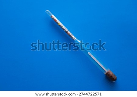 ​A glass hydrometer with a colored scale is positioned diagonally on a deep blue background. This laboratory device provides accurate measurement of liquid density and alcohol strength,