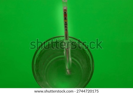 A glass hydrometer is immersed in a clear liquid inside a glass against a bright green background. This laboratory device is used to measure the density of alcohol or other solutions, making the shot relevant to home brewing, chemistry, and quality control.