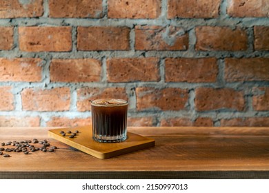 A Glass of ice coffee, cold americano in the morning over a red bricks wall background, and coffee beans on wooden counter bar table. Cold brew refreshment summer drink with copy space. Cafe Concept.