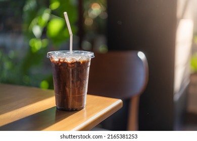 Glass Of Iced Coffee With Milk, Chocolate Syrup In Coffee Shop. Cold Mocha Coffee In Plastic Cup On Wooden Table In Cafe With Plant And Sunlight Background