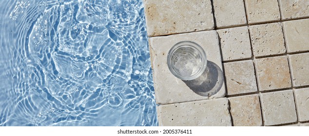 A glass of iced cold water placing on the travertine marble stone by the beautiful swimming pool in the sunny morning.