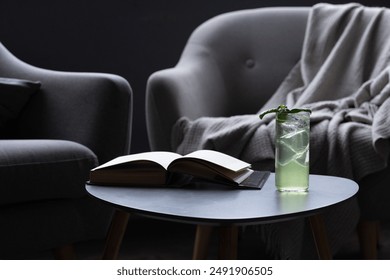 Glass of iced drink with sprig of mint on round table next to book. Gray armchair with throw blanket behind table. Room is dimly lit. Copy space, place for product display - Powered by Shutterstock