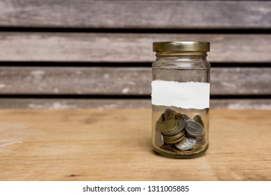 Glass jar filled with coins attached with empty white paper on wooden background. Free copy space. Financial concept.