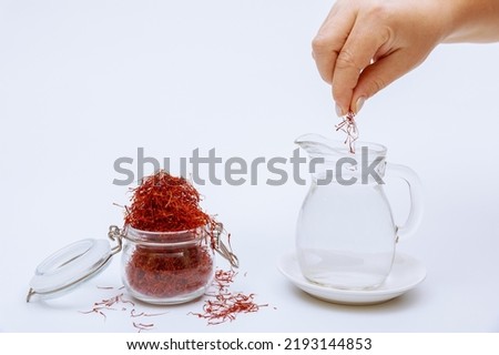 A glass jar filled with saffron stamens and a woman putting red stamens into water in a decanter on a white background. Dry red spice.