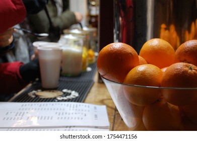 Glass Jar Full Of Oranges In Foreground On A Bar Counter And Some People's Hands Wearing Gloves Aside Holding Hot Drinks At Budapest Food Market In Winter Time