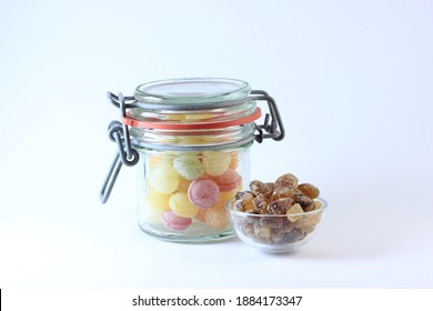 Glass Jar With Home Made Yellow, Red, Orange, Green Sugar Bonbon Sweets With Smaller Glass Jar Containing Brown Rock Candy Sugar, White Background