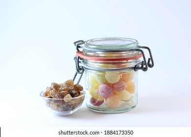 Glass Jar With Home Made Yellow, Red, Orange, Green Sugar Bonbon Sweets With Smaller Glass Jar Containing Brown Rock Candy Sugar, White Background