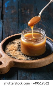 Glass Jar Of Homemade Salted Caramel Sauce On Wooden Serving Board With Ingredients Brown Sugar Above. Caramel Flowing From Spoon. Over Old Dark Blue Wooden Background.