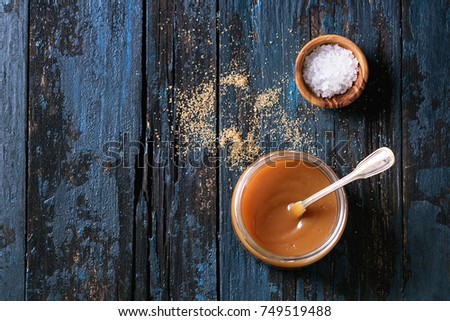 Glass jar of homemade salted caramel sauce with spoon, brown sugar and bowl of salt. Over old dark blue wooden background. Top view with space