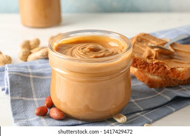 Glass jar with peanut butter, peanut, kitchen towel, spoon and peanut butter sandwich on white wooden background, space for text and closeup