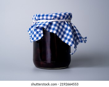 Glass Jar With Red Jam Decorated With Blue And White Checkered Cloth On Gray Studio Background. Homemade Canned Raspberry, Strawberry Or Cherry Jelly. Sweet Dessert Made From Fruits Or Berries.