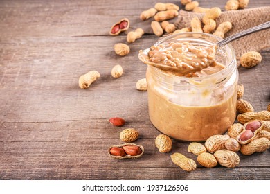 Glass jar and spoon full of crunchy peanut butter with peanuts in nutshell nearby on wooden background. Copy space.