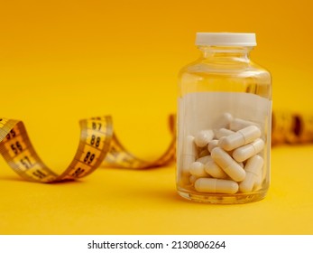 glass jar with tablets in capsules stands in front of a measuring tape on a yellow background, close-up. The concept of weight loss, fat burner, vitamins, sports nutrition.