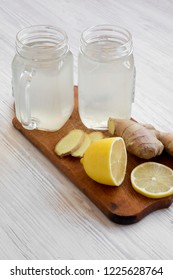 Glass jars of ginger tea with ingredients on wooden board over white wooden surface, side view. Close-up.