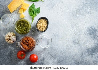 Glass jars with red and green pesto and cooking italian recipe ingredients Parmesan cheese, basil leaves, pine nuts, olive oil, garlic, salt, tomatoes top view on grey concrete surface space for text - Powered by Shutterstock