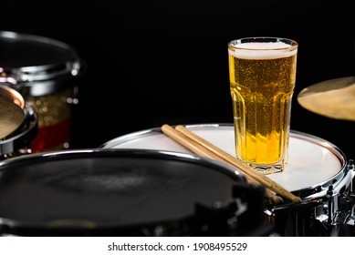 Glass Of Light Beer On Professional Drum Set Closeup. Drumsticks, Drums And Cymbals, At Live Music Rock Concert, In The Club Stage, Bar, Or In Recording Studio. Black Background.