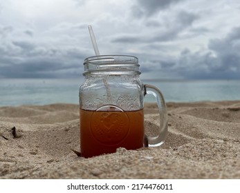 A Glass Mason Jar With Rum Punch On A Beach With Dramatic Clouds And The Ocean In The Background