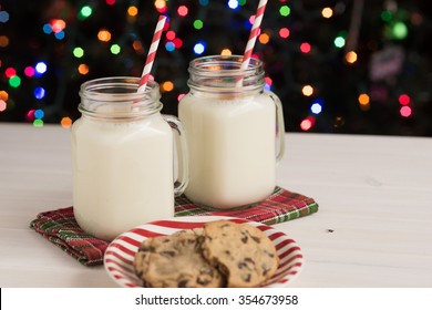 Glass Mason Jars Filled With Homemade Egg Nog With Cinnamon And Red Swirled Straws And Chocolate Chip Cookies On Red And White Striped Plate And Christmas Tree Lights In Background