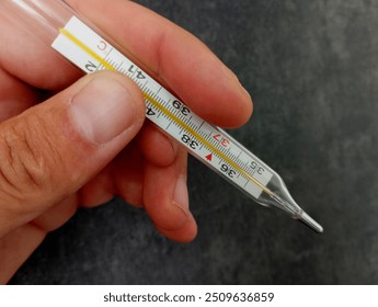 A glass mercury thermometer for measuring body temperature in a human hand on a black textured background. Medical thermometer.