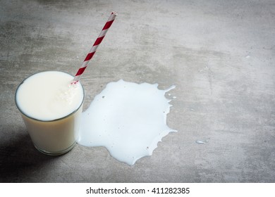 Glass Of Milk And A Red And White Drinking Straw On A Concrete Table. It Is A Puddle Of Milk On The Table. Crying Over Spilt Milk.