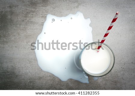 Glass of milk and a red and white drinking straw on a concrete table. It is a puddle of milk on the table. Crying over spilt milk. View from the above.