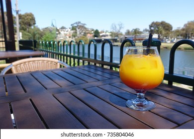 A Glass Of Orange Juice Mocktail On A Wood Table By A River In A Sunny Weather. City Of Maribyrnong, Melbourne.