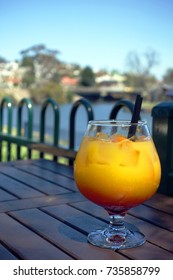 A Glass Of Orange Juice Mocktail On A Wood Table With A Blurry River View In A Sunny Weather. City Of Maribyrnong,