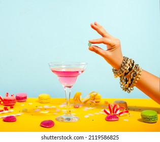 Glass Of Pink Cocktail With Heart Candies And Macaroons. Birthday Party Or Valentines Day Concept. Woman Hand Put Hard Candy In A Glass.