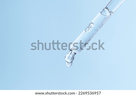 Glass pipette with facial serum with falling drop on blue background. Liquid skin care cosmetic product with bubbles closeup. Essential oil droplet macro. Hyaluronic acid skin treatment in dropper.