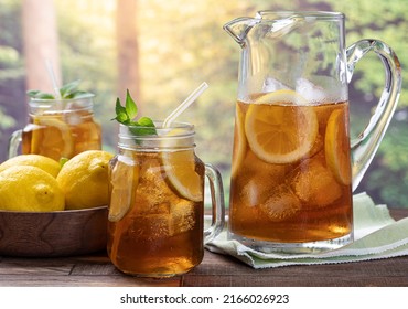 Glass and pitcher of iced tea with mint, lemon slices and ice on an outdoor wooden table with rural summer background