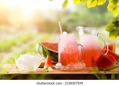 Glass and pitcher with natural watermelon drink with a lot of ice on a wooden table with fruit around and a nature background with orchard. Front view. Horizontal composition.