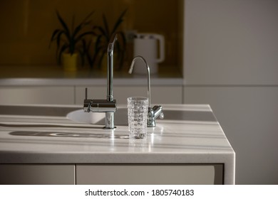 Glass Of Pure Filtered Water On The Kitchen Table In The Rays Of Sunlight. Kitchen Faucet And Reverse Osmosis Faucet For Water Purification. Healthy Lifestyle Concept. Сlose-up Shot, Selective Focus.