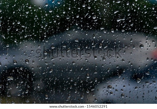 A glass with rain drops that have a backdrop as a
car in a parking lot.