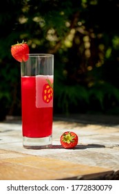 Glass With Red Fruit Drink And Strawberry Image And Garnish On An Outdoor Tiled Tabletop Against A Green Pinetree Background