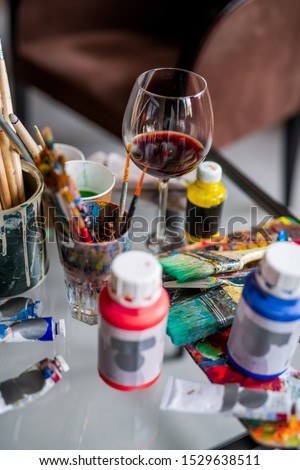 Glass of red wine among paintbrushes and various paints and gouaches on workplace of an artist