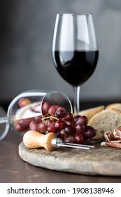 A Glass Of Red Wine With Another Wine Glass Tipped Over With Red Grapes Spilling Out On A Rustic Board.