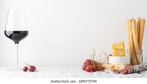 Glass of red wine and cheese, sausage, grapes and grissini bread sticks on the table. Light background, traditional wine snacks on tablecloth covered table - Powered by Shutterstock