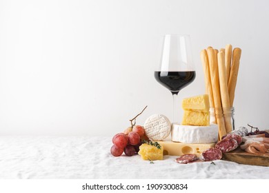 Glass of red wine and cheese, sausage, grapes and grissini bread sticks on the table. Light background, traditional wine snacks on tablecloth covered table - Powered by Shutterstock