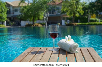 Glass Of Red Wine And A Cotton Towel On A Sunbed Near A Swimming Pool In A Late Morning Of Summer, Village House In Background.