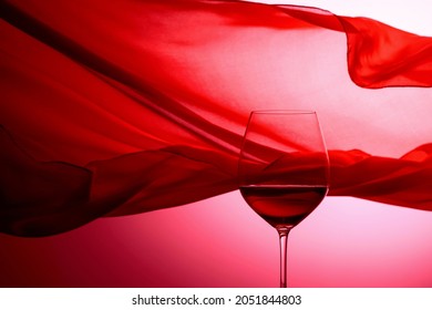 Glass Of Red Wine On A Background Of Waving Red Satin Curtain.