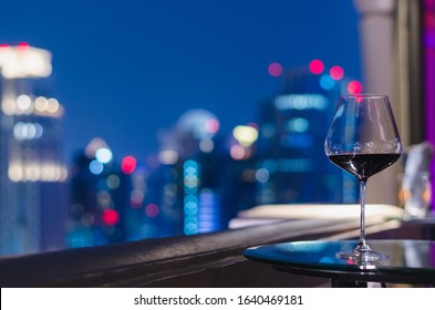 A Glass Of Red Wine On Table Of Rooftop Bar With Colorful Bokeh Of City Lights.