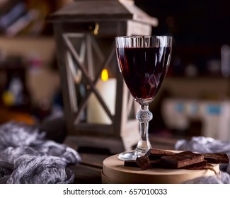 Glass Of Red Wine On The Table. Wooden Lantern In The Background. The Candle Burns In The Lantern. Dark Background. Chunks Of Chocolate Nearby. The Atmosphere Of Evening And Comfort