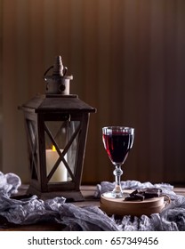 Glass Of Red Wine On The Table. Wooden Lantern In The Background. The Candle Burns In The Lantern. Dark Background. Chunks Of Chocolate Nearby. The Atmosphere Of Evening And Comfort