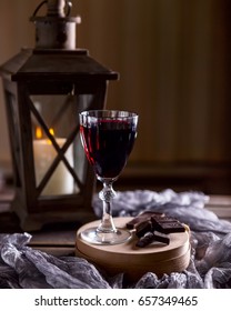 Glass Of Red Wine On The Table. Wooden Lantern In The Background. The Candle Burns In The Lantern. Dark Background. Chunks Of Chocolate Nearby. The Atmosphere Of Evening And Comfort