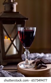 Glass Of Red Wine On The Table. Wooden Lantern In The Background. The Candle Burns In The Lantern. Dark Background. Chunks Of Chocolate Nearby. The Atmosphere Of Evening And Comfort