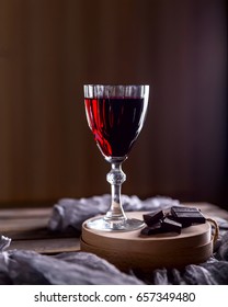 Glass Of Red Wine On The Table. Dark Background. Chunks Of Chocolate Nearby. The Atmosphere Of Evening And Comfort