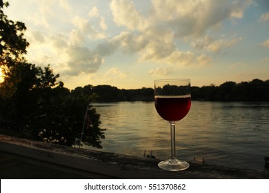 Glass Of Red Wine Rests On A Deck Railing At Dusk With A Lake And Treeline And Clouds In The Background