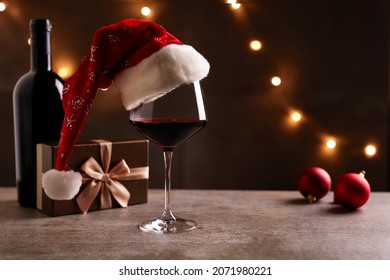 Glass Of Red Wine With Santa Claus Hat, Bronze Gift Box And Christmas Decorations On Table. Christmas Lights In The Black Background.