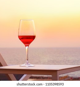 A Glass Of Red Wine Standing On A Table Near A Summer Chair Against A Background Of A Sea Horizon At Sunset. A Glass Of Red Wine Against The Background Of The Evening Sea Horizon At Sunset.